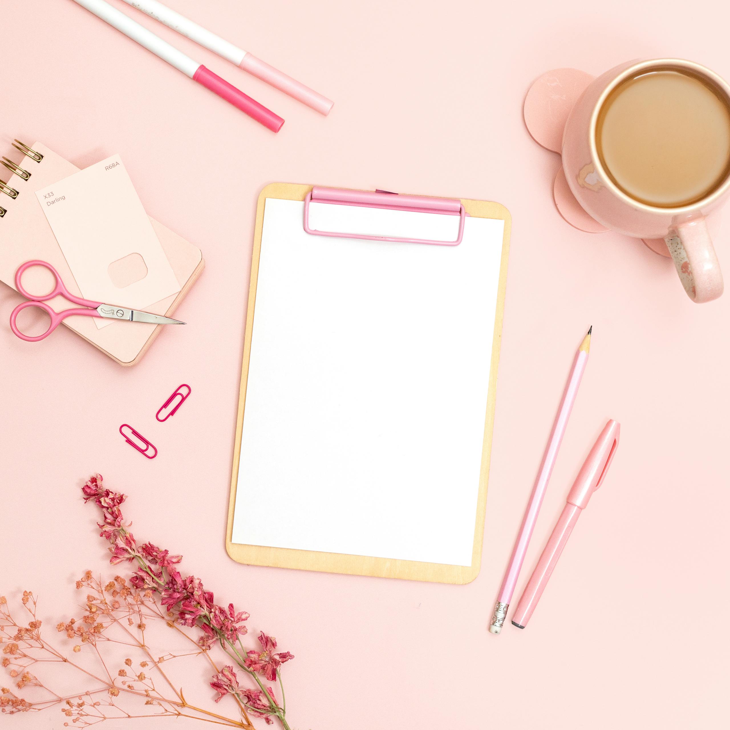 Minimalist flat lay of pink office supplies featuring a clipboard, stationery, and coffee cup.